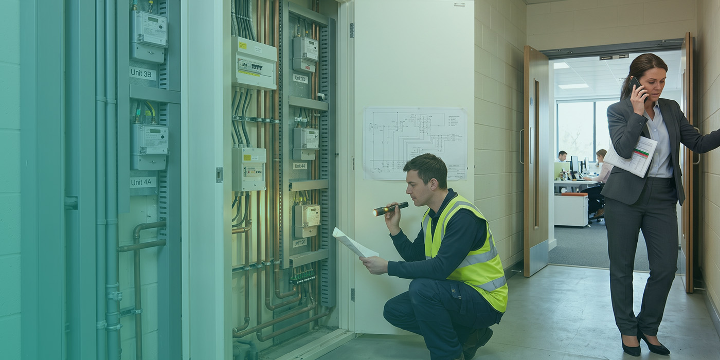 A utilities engineer in a yellow high-visibility vest crouching to inspect a multi-tenant electrical metering cabinet with a torch and schematic drawing, whilst a building manager walks past on a phone call holding documents, with an occupied office visible through an open doorway and an electrical schematic pinned to the wall