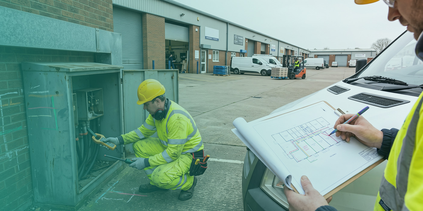 Two utilities operatives on an active industrial estate, one crouching to test an external supply cabinet with a clamp meter, the other annotating a colour-coded electrical site layout drawing resting on a van bonnet