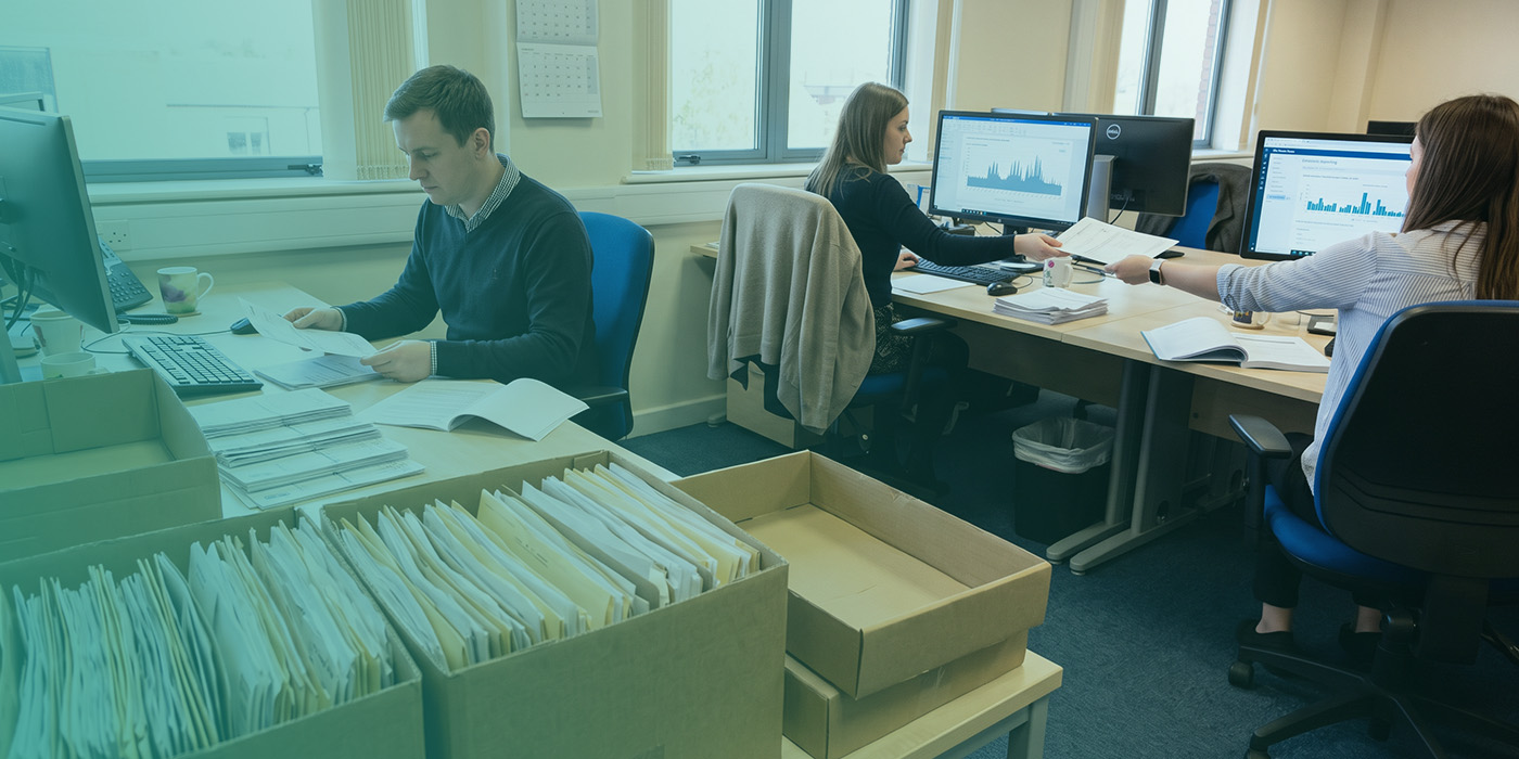 Three utilities billing analysts working at adjacent desks in a busy back-office environment, one reviewing paper invoices against an open ledger, a second working at a monitor showing a consumption spike chart, and a third passing a printed document across to a colleague, with two archive boxes of filing in the foreground
